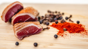 Slices and chubs of il porcellino salumi's Duck Prosciutto on a cutting board with herbs and spices next to them.