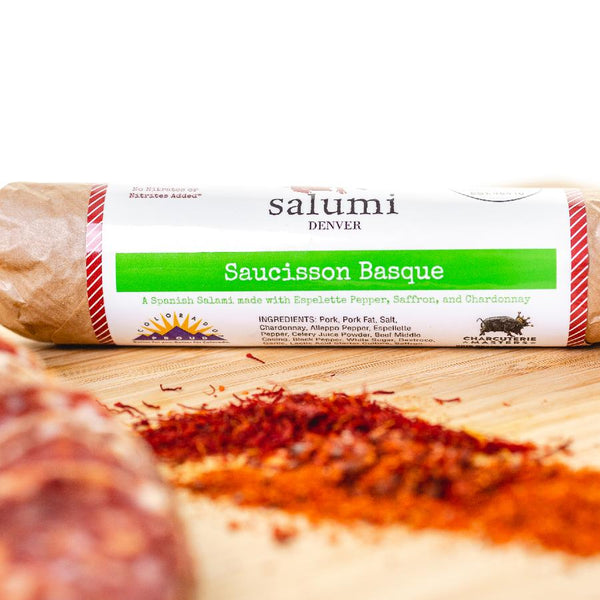 A close up of Saucisson Basque salami in packaging on a cutting board with blurred salami slices, herbs and spices in the foreground.