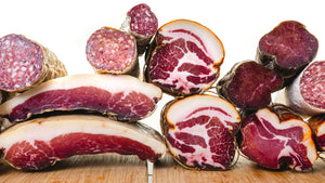 A picture looking at the cross section of several types of cured meat like salami, coppa, speck, spanish lomo and more. All the meat is stacked on top of each other and top of cutting boards.