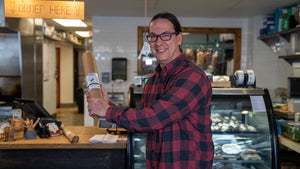 A picture of il porcellino salumi's owner, Bill Miner, holding the large format Black Truffle Salami like a baseball bat at his deli with the meat case, drying room, and cash register in the background.