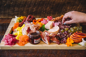 A colorful charcuterie board filled with meat, cheese, vegetables, fruits and a jar of jam in the front. The charcuterie board is on a wood table and a hand grabbing a piece of meat off the board enters the image from the right.
