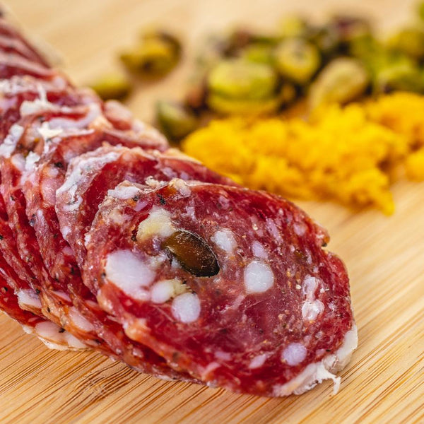 A close up of slices of hard salami with pistachios, fat, herbs and spices in it. The salami is on a cutting board with blurred pistachios and orange zest in the background.
