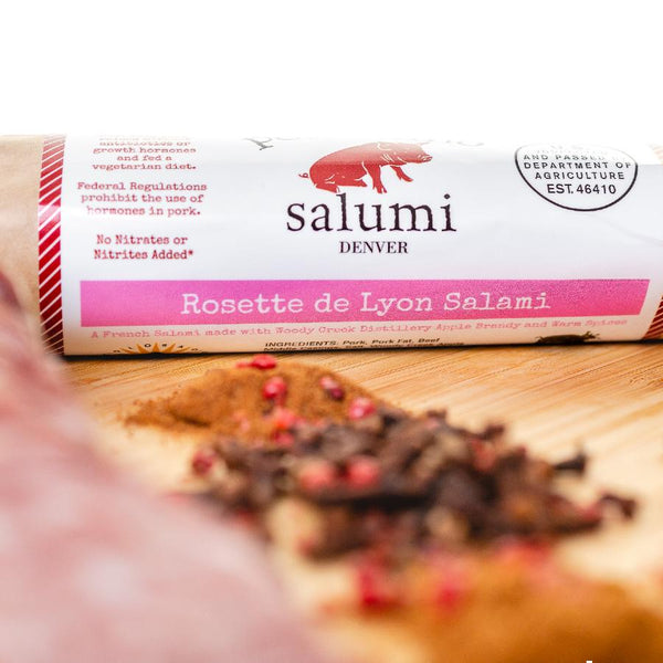 A close up of Rosette de Lyon salami in packaging on a cutting board with blurred herbs and spices in the foreground.